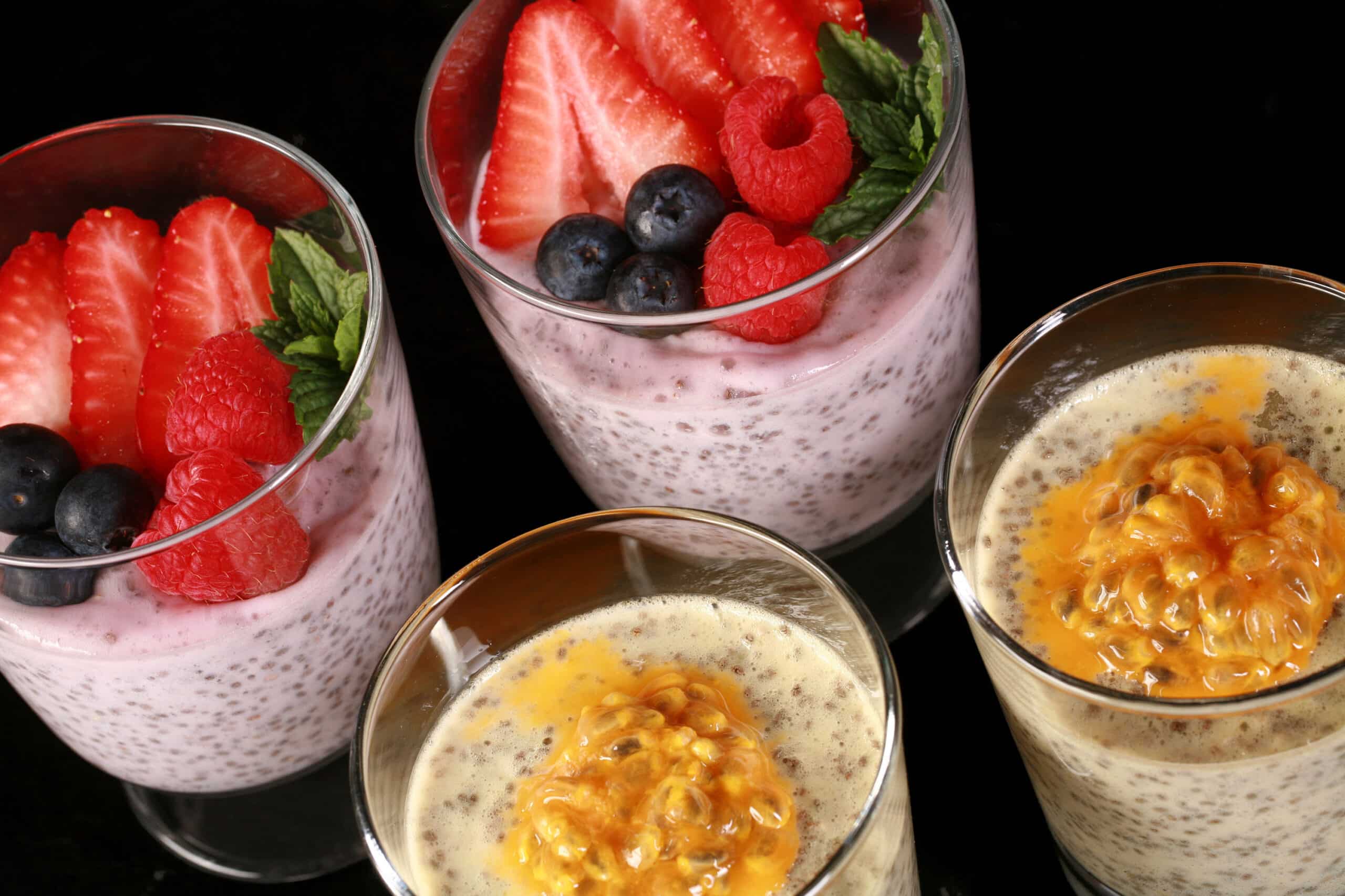 4 glasses of keto protein pudding with chia seeds. 2 are yellow and topped with passionfruit, the other two are pink and topped with berries.