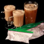 2 glass mugs of keto cappuccino and a tall glass of protein iced coffee, with several bags of protein powder cappuccino mix.