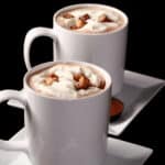 2 mugs of protein peanut butter hot chocolate with whipped cream and chopped peanut butter cups.