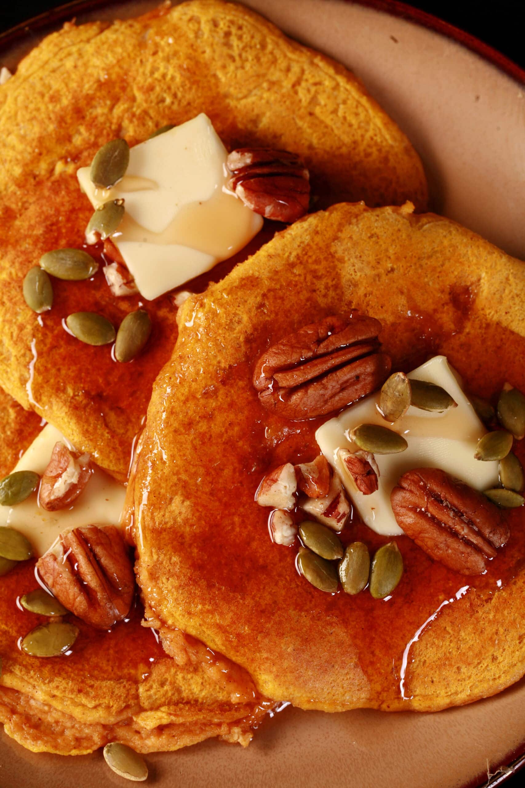 3 low carb pumpkin protein powder pancakes on a plate, topped with butter, syrup, pecans, and pumpkin seeds.