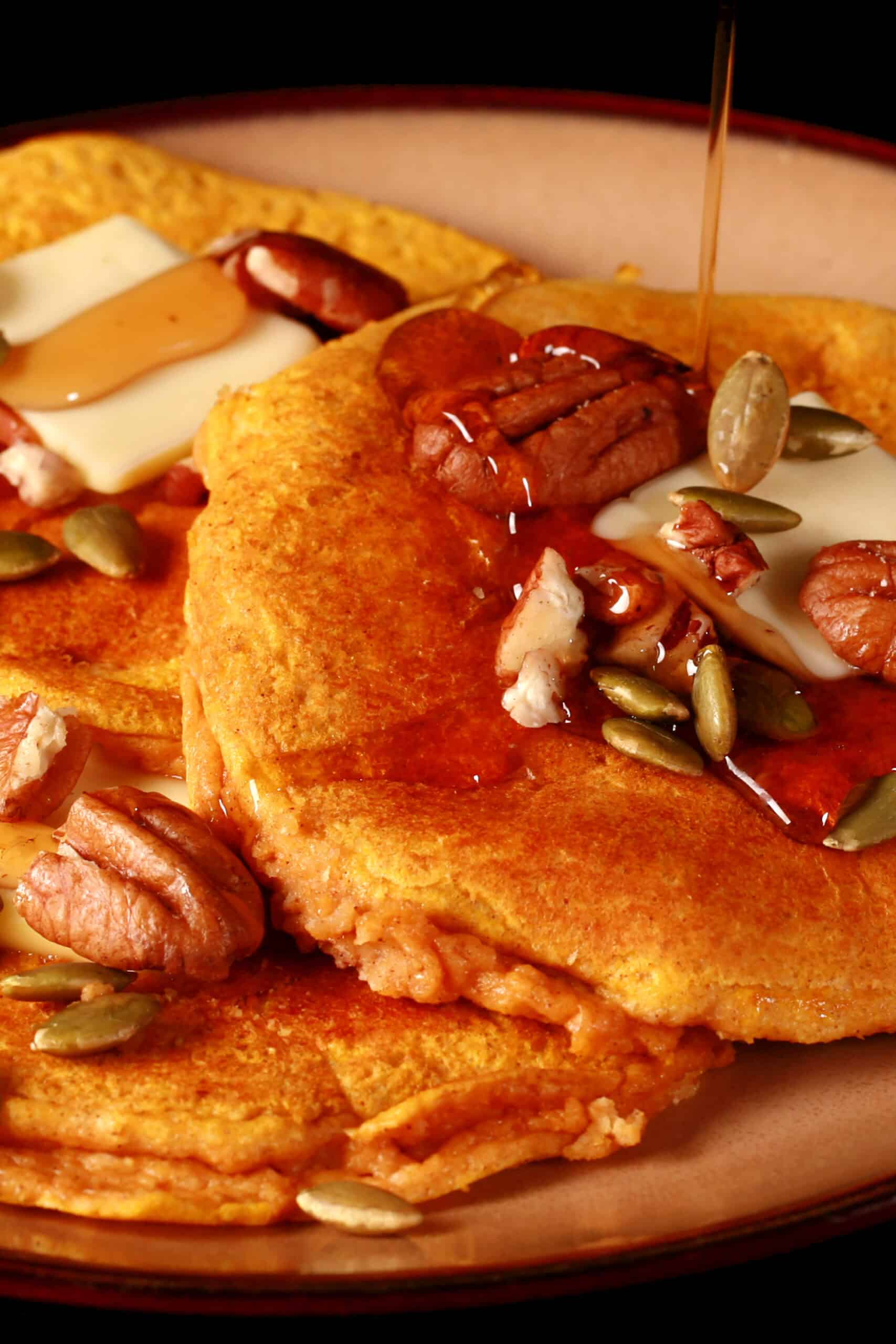 3 low carb pumpkin protein powder pancakes on a plate, topped with butter, syrup, pecans, and pumpkin seeds.