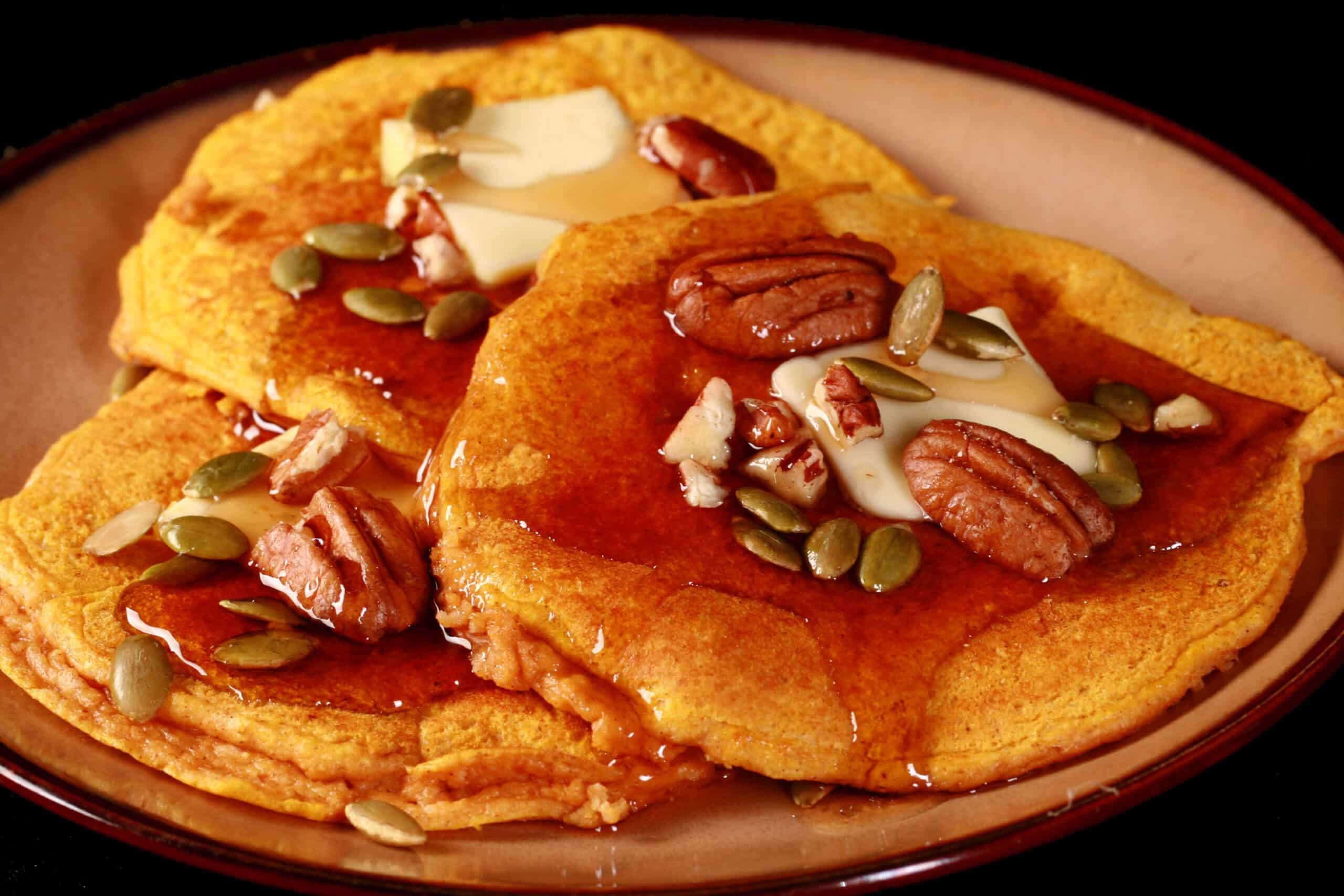 3 low carb pumpkin protein pancakes on a plate, topped with butter, syrup, pecans, and pumpkin seeds.