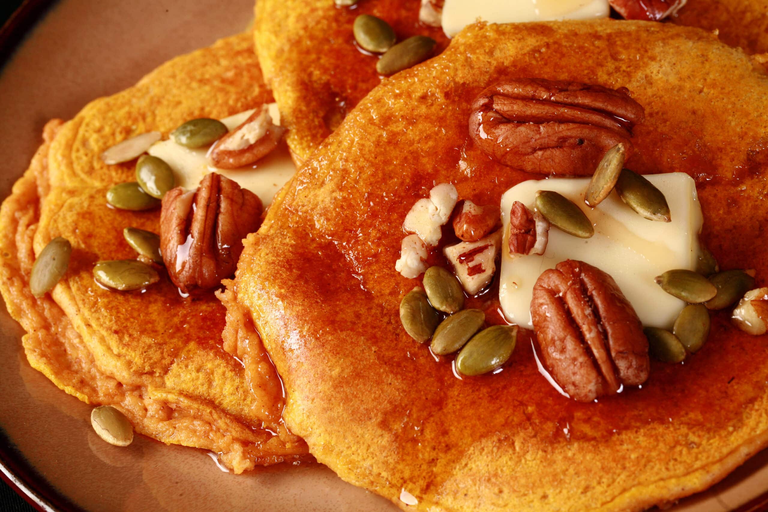 3 keto pumpkin protein pancakes on a plate, topped with butter, syrup, pecans, and pumpkin seeds.