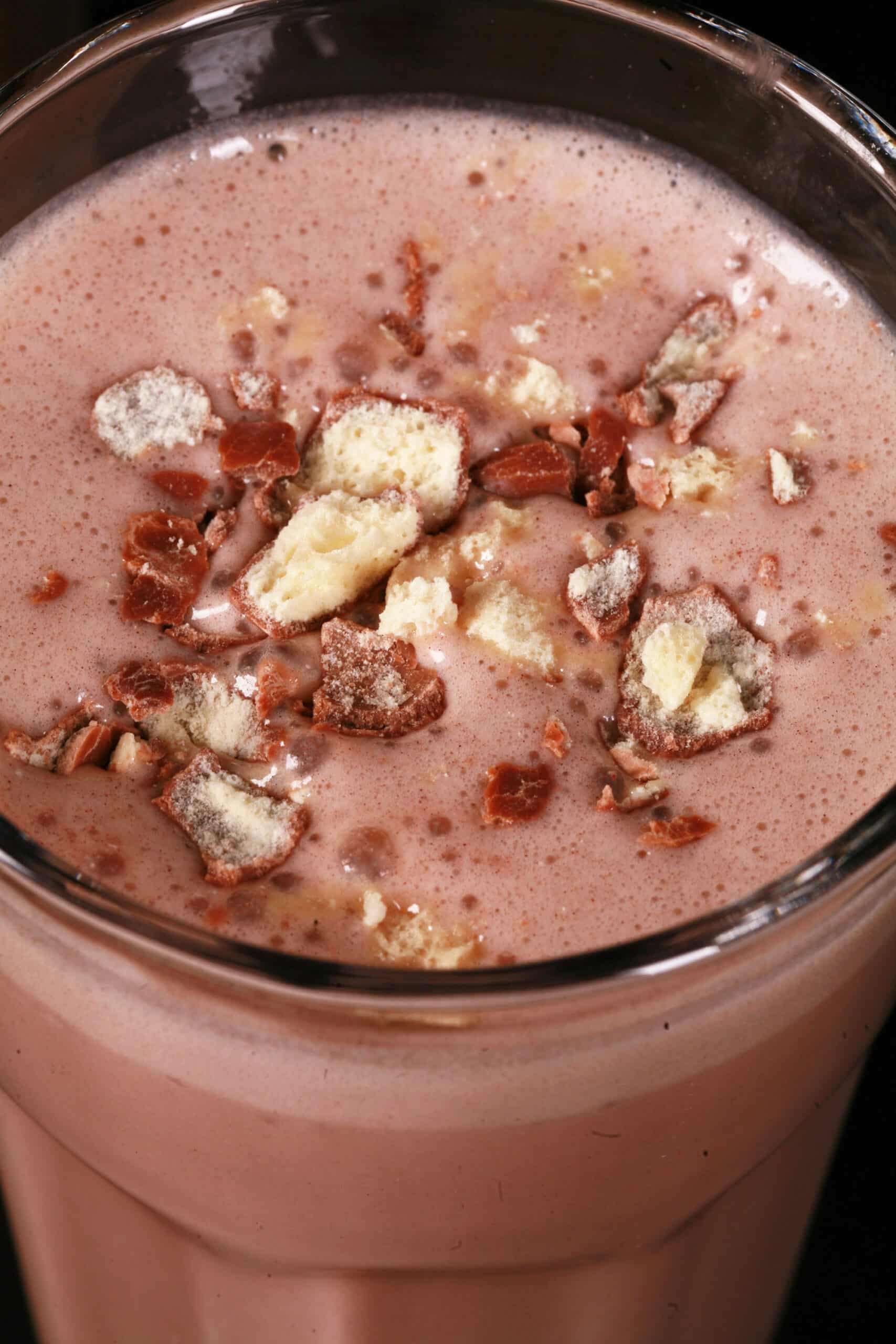 A tall glass of chocolate malted milk protein shake with crushed Whoppers Malted milk balls candy on top.