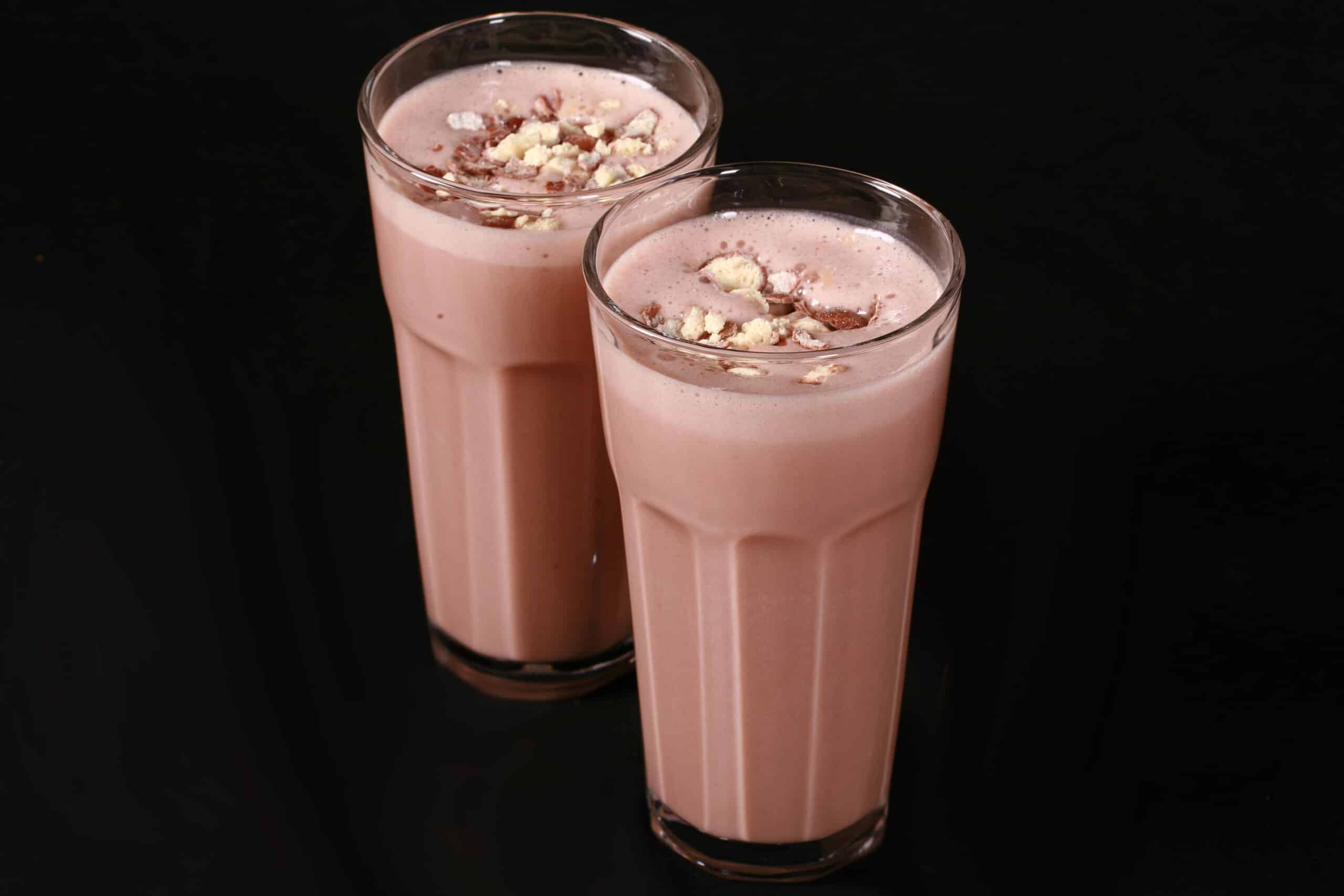 2 tall glasses of chocolate malt protein shakes with crushed Whoppers Malted milk balls candy on top.