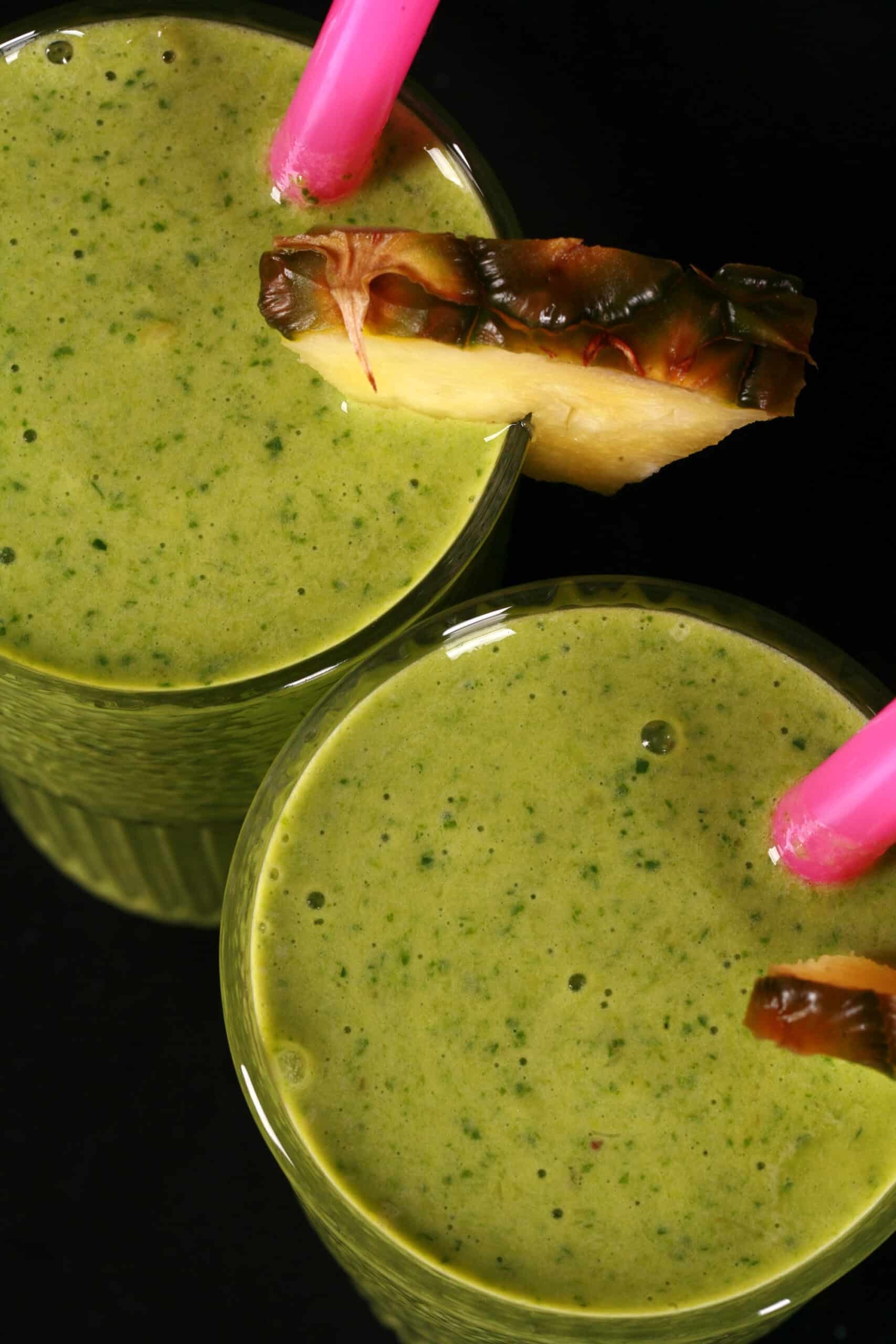 2 tall glasses of tropical green protein smoothies, garnished with fresh pineapple wedges and pink straws.