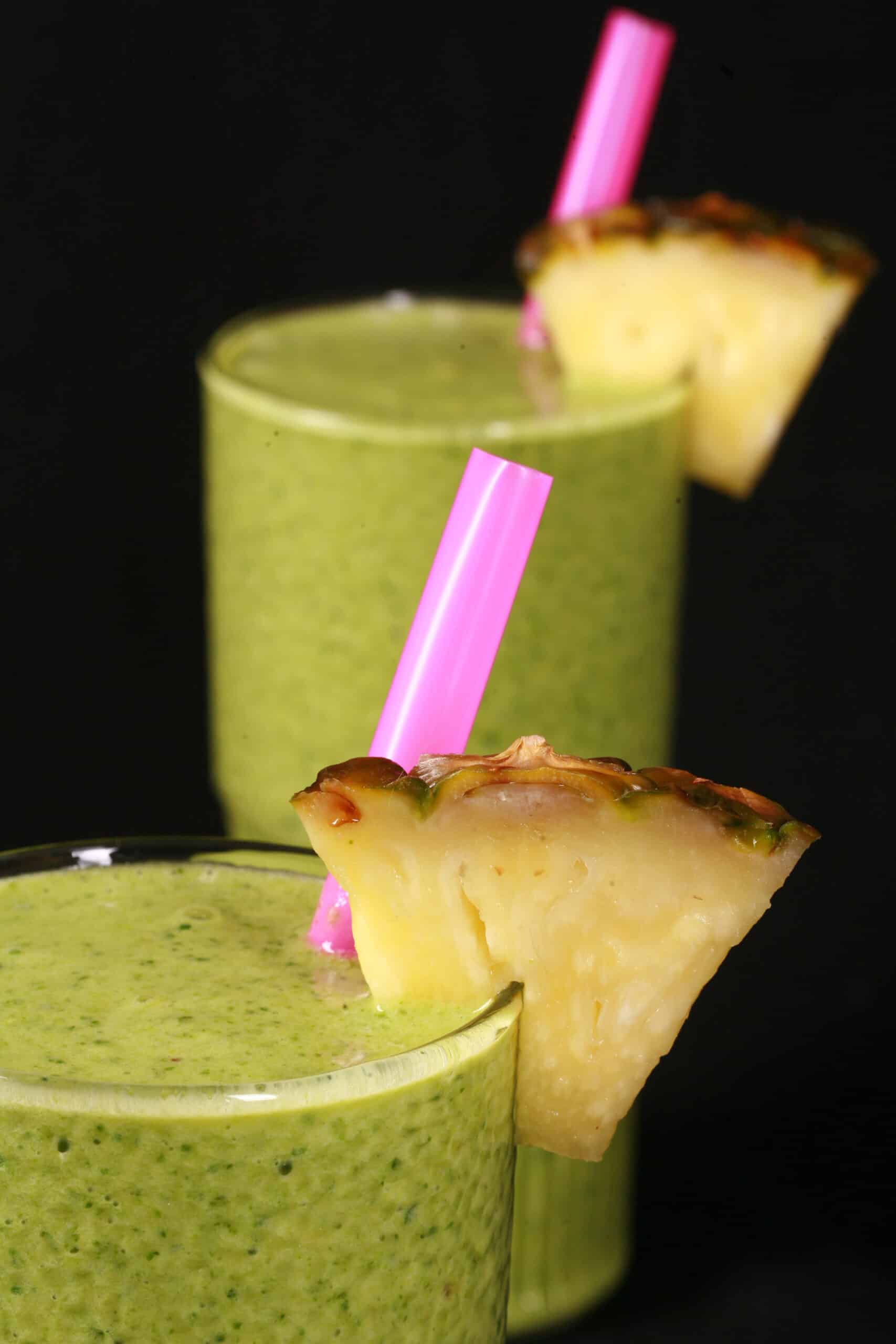 2 tall glasses of high protein tropical green smoothies, garnished with fresh pineapple wedges and pink straws.