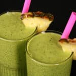 2 tall glasses of tropical green protein smoothies, garnished with fresh pineapple wedges and pink straws.