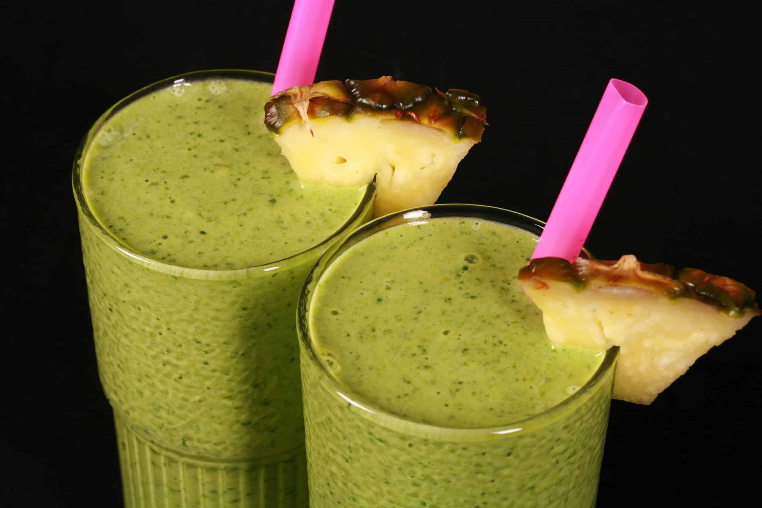 2 tall glasses of tropical green protein smoothies, garnished with fresh pineapple wedges and pink straws.