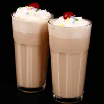 2 tall glasses of high protein malted milk smoothies garnished with whipped cream, sprinkles, and a maraschino cherry.