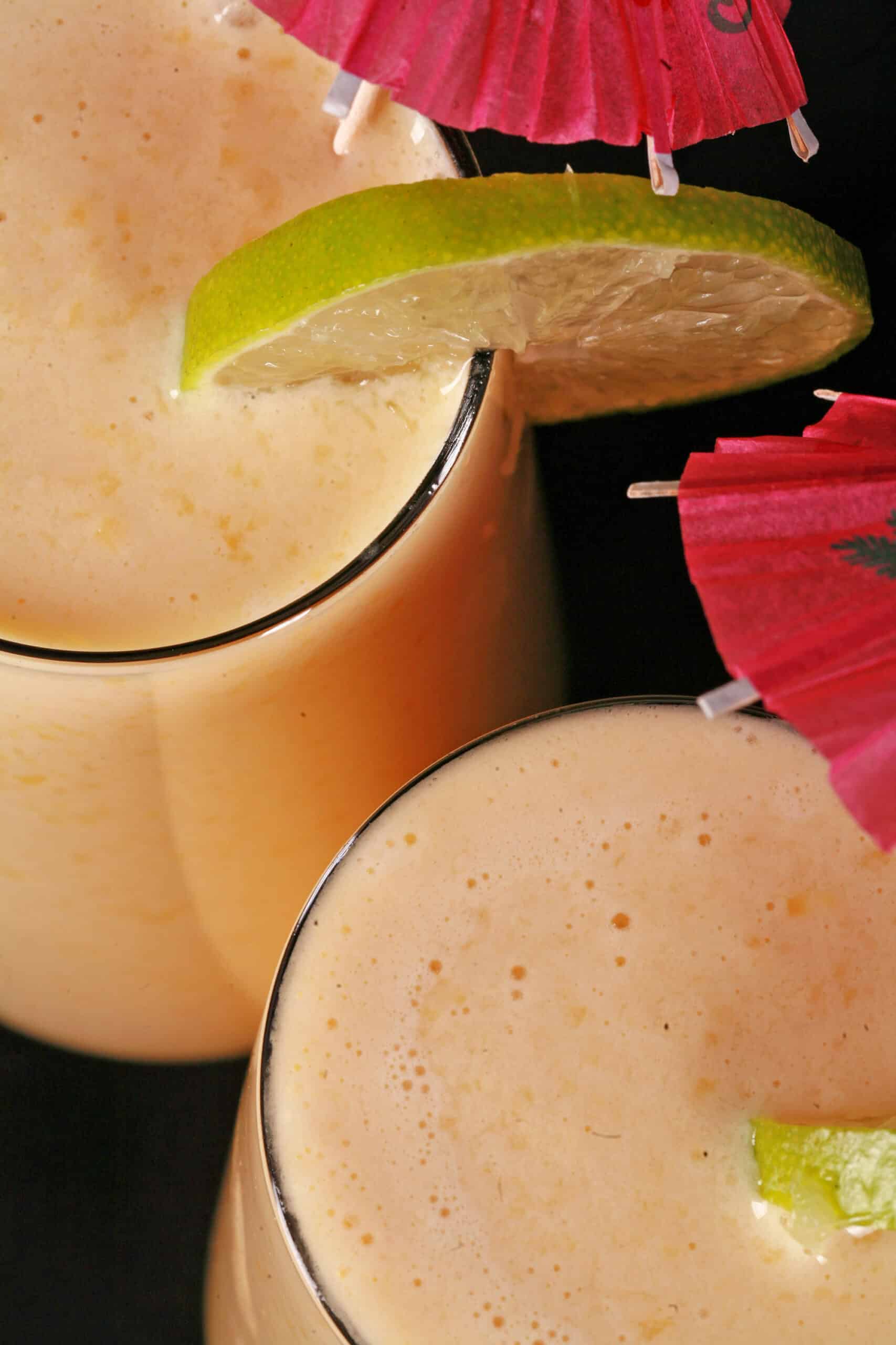 2 tall mango daiquiri protein smoothies, garnished with lime slices and pink cocktail umbrellas.