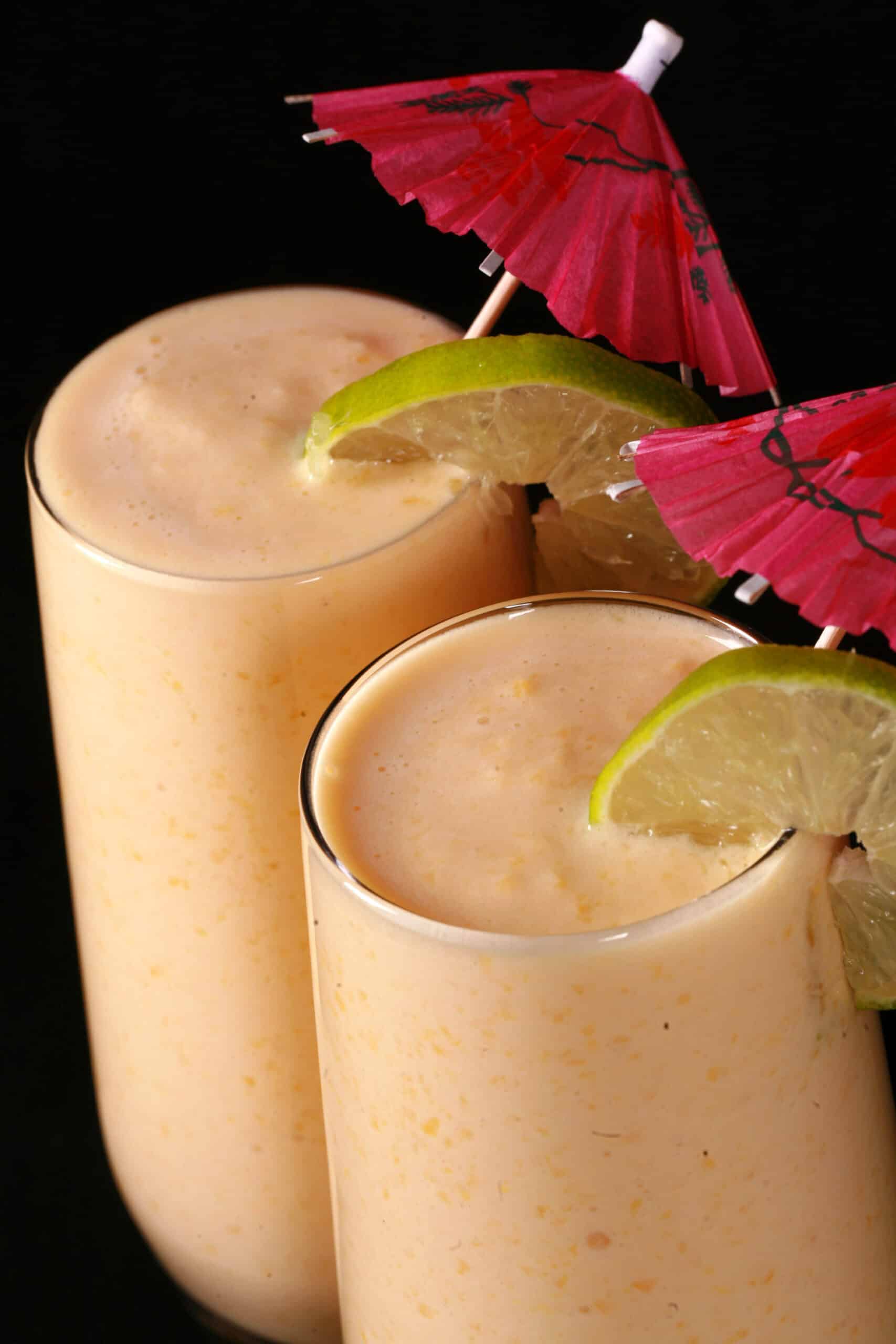 2 tall glasses of mango daiquiri protein smoothie, garnished with lime slices and pink cocktail umbrellas.