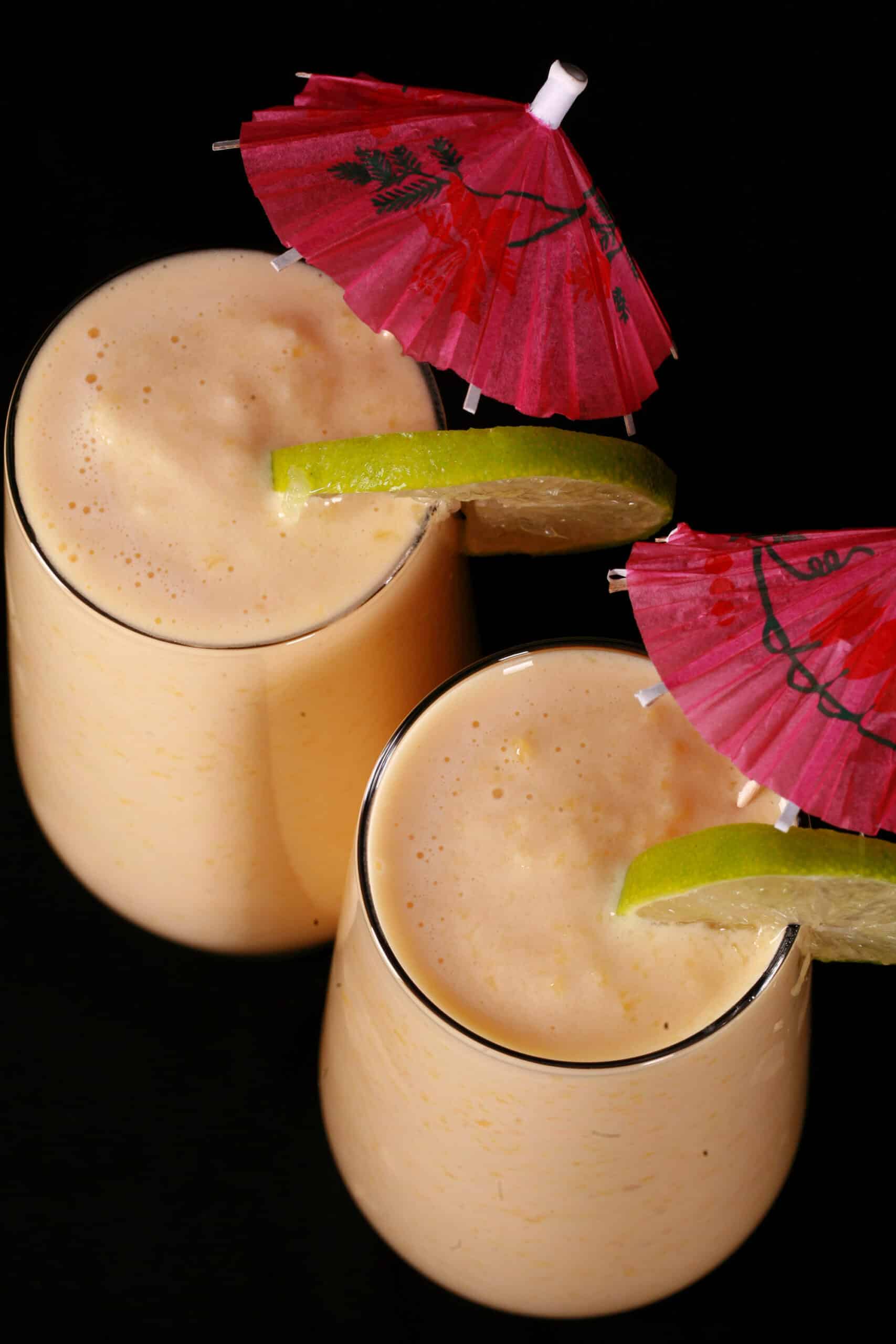 2 tall mango daiquiri protein smoothies, garnished with lime slices and pink cocktail umbrellas.