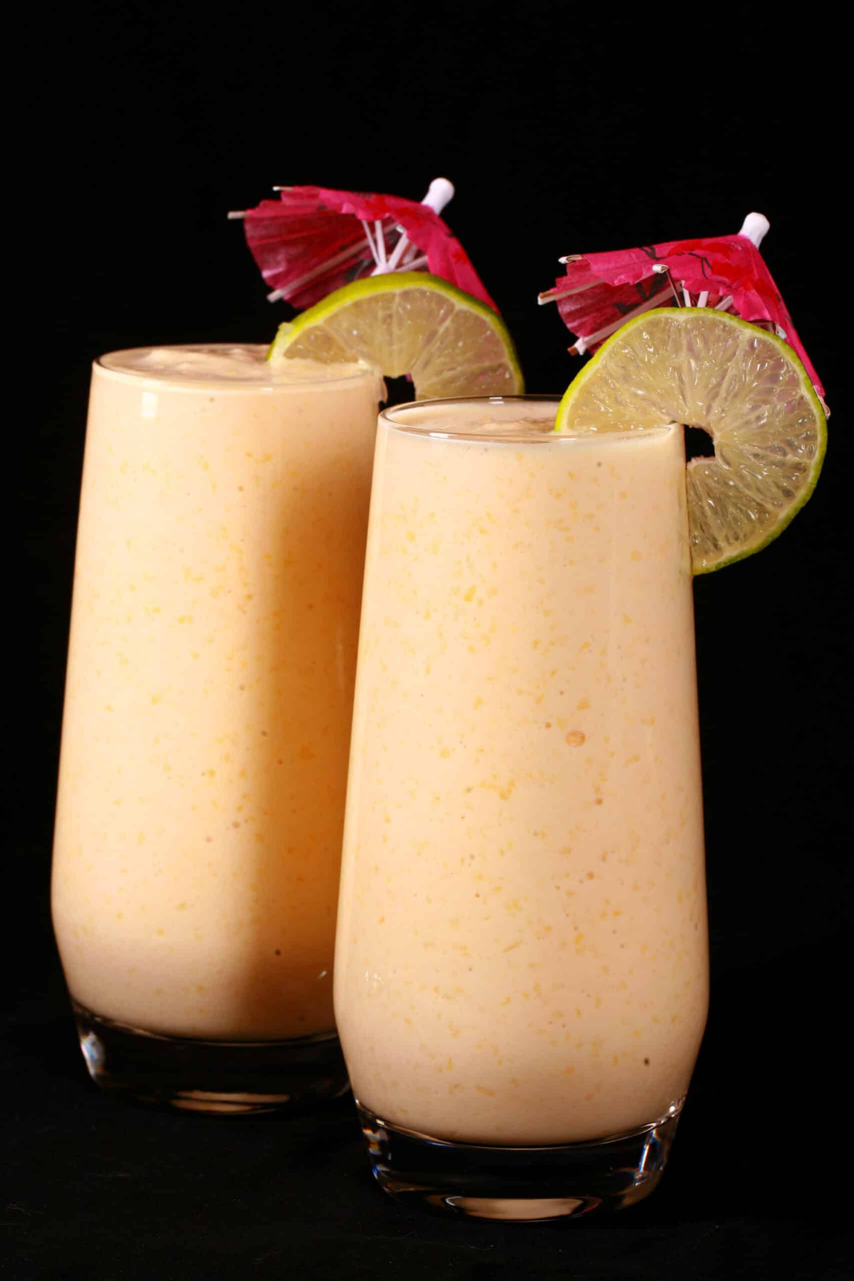 2 tall mango daiquiri protein smoothies, garnished with lime slices and pink cocktail umbrellas.