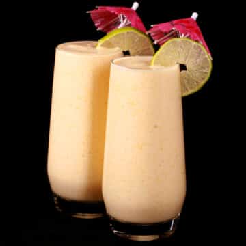 2 tall glasses of mango daiquiri protein smoothie, garnished with lime slices and pink cocktail umbrellas.