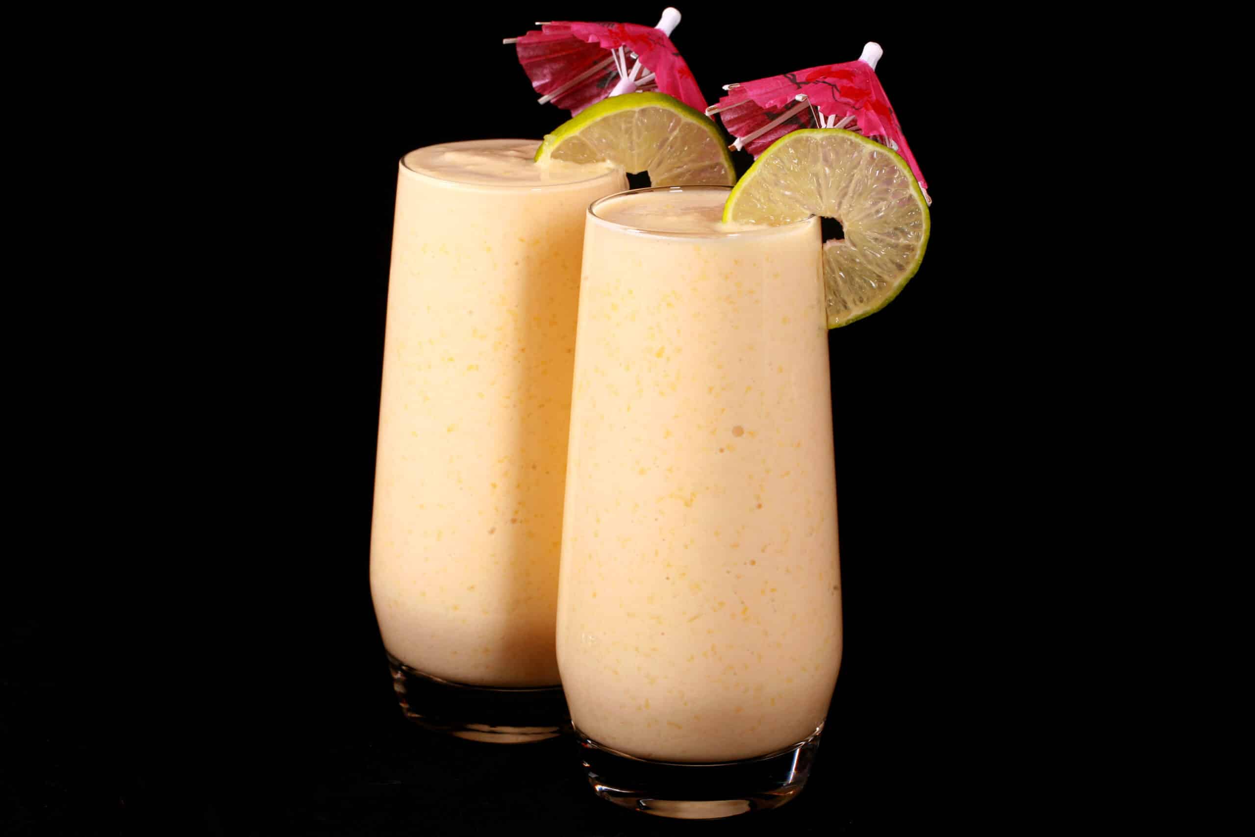 2 tall glasses of mango daiquiri protein smoothie, garnished with lime slices and pink cocktail umbrellas.