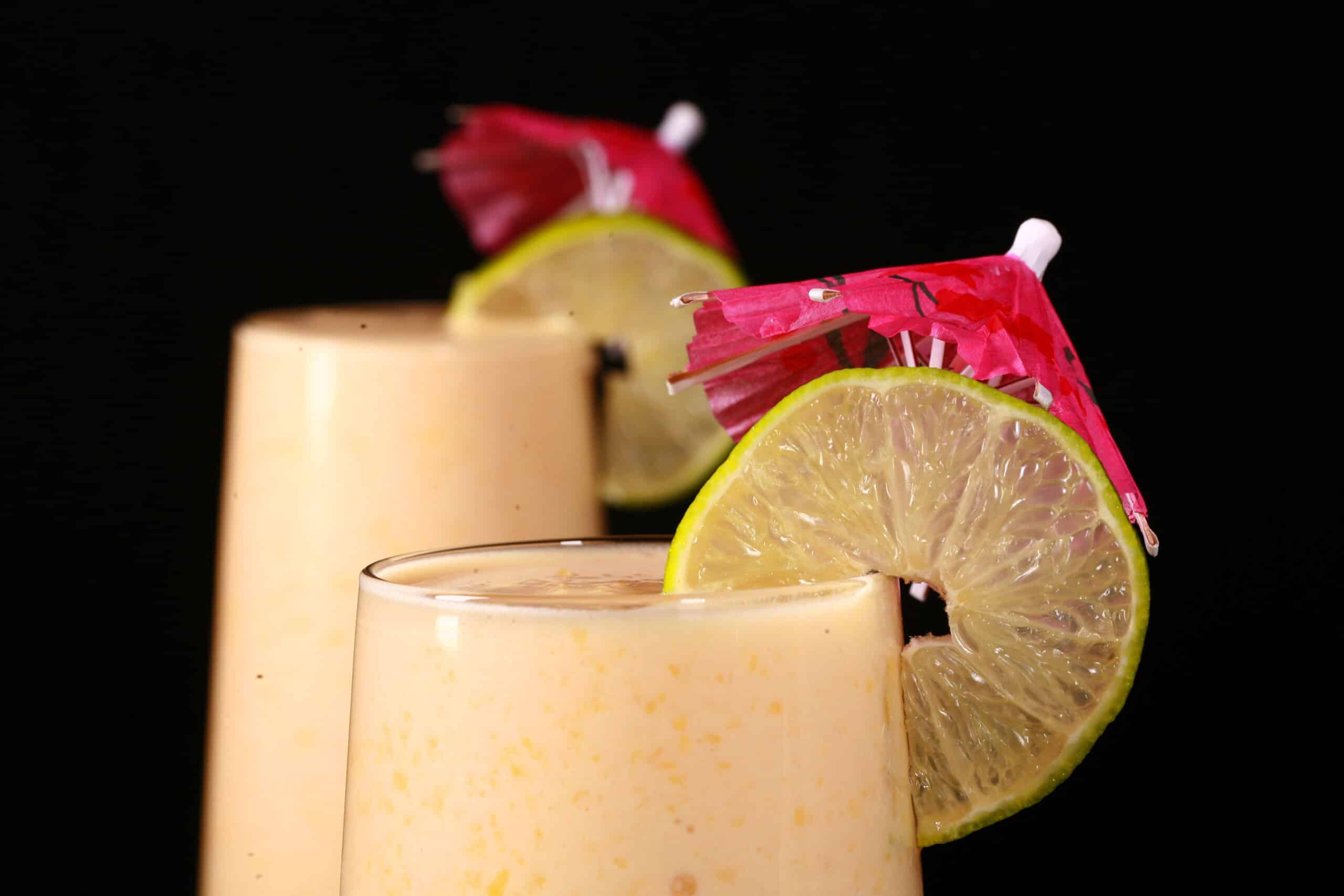 2 tall mango daiquiri protein smoothies, garnished with lime slices and pink cocktail umbrellas.