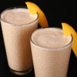 2 tall glasses of mango lassi protein smoothies, garnished with slices of fresh mango.