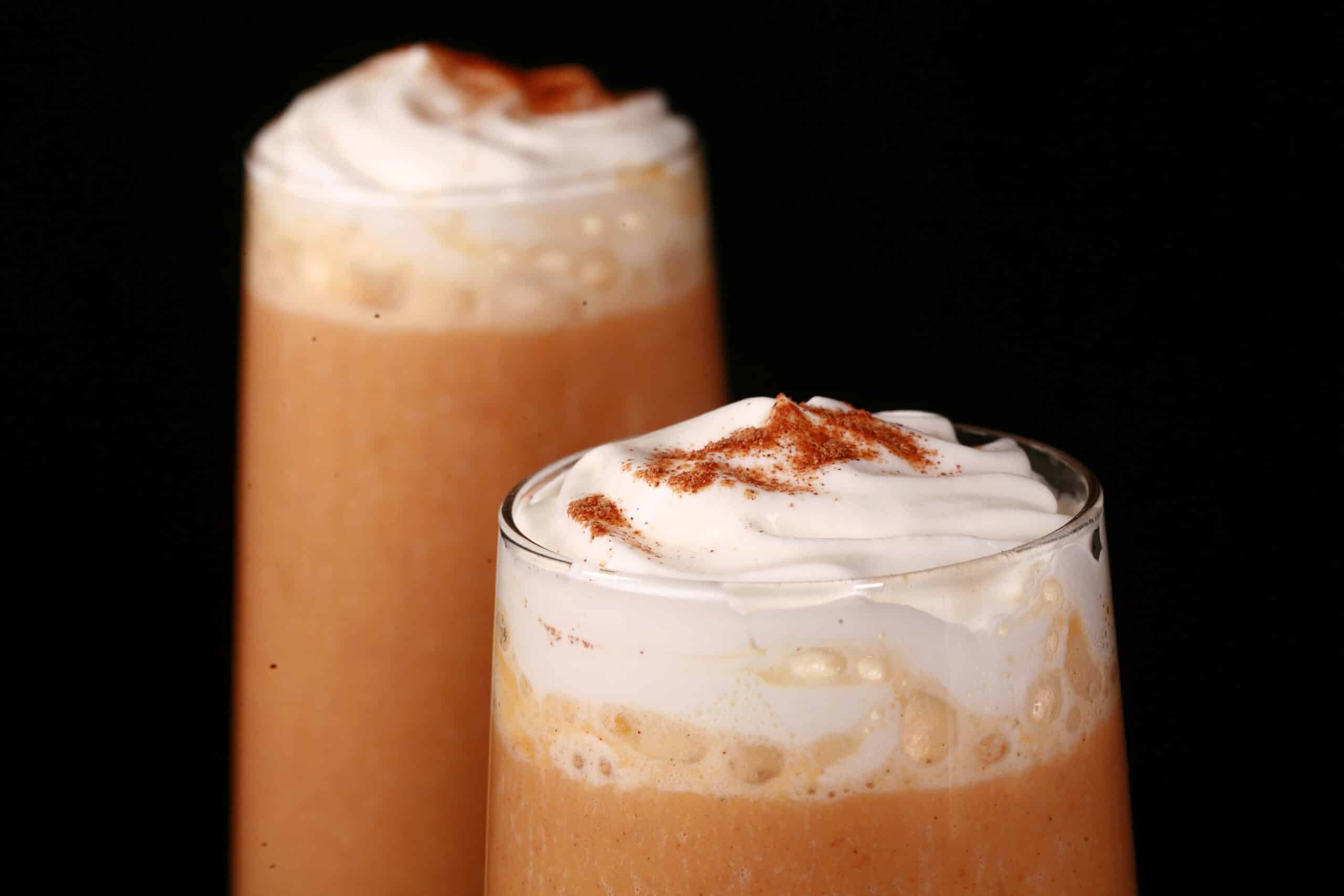 2 pumpkin pie protein smoothies in tall glasses, topped with whipped cream and pumpkin spice.