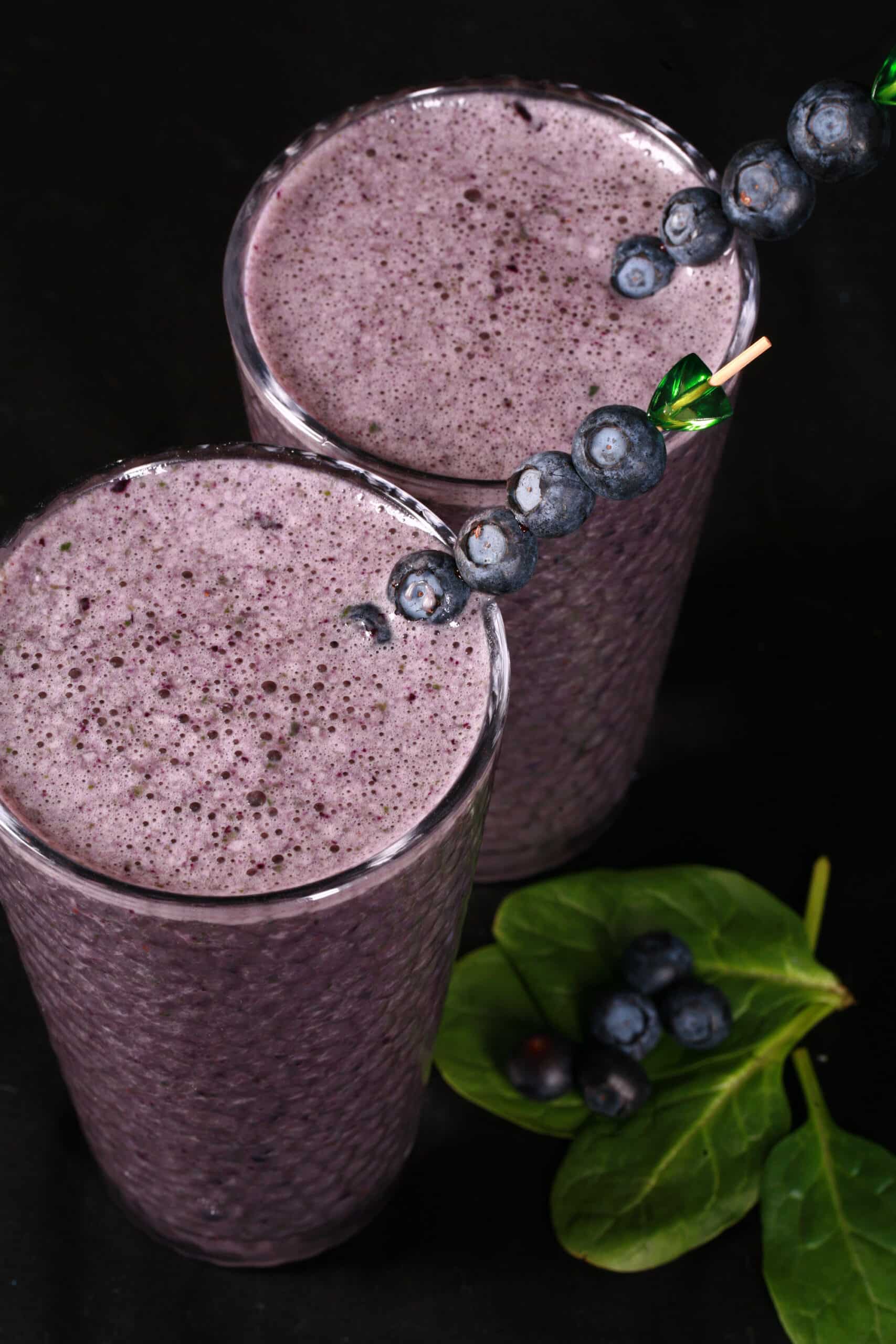 2 tall glasses of high protein blueberry spinach smoothies, garnished with fresh blueberries and baby spinach.