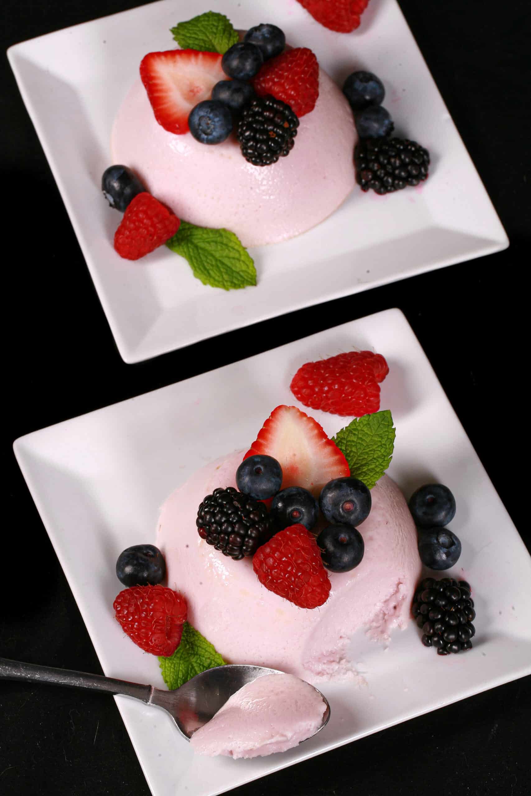 2 light pink high protein panna cotta servings on plates, with mixed berries and mint leaves.
