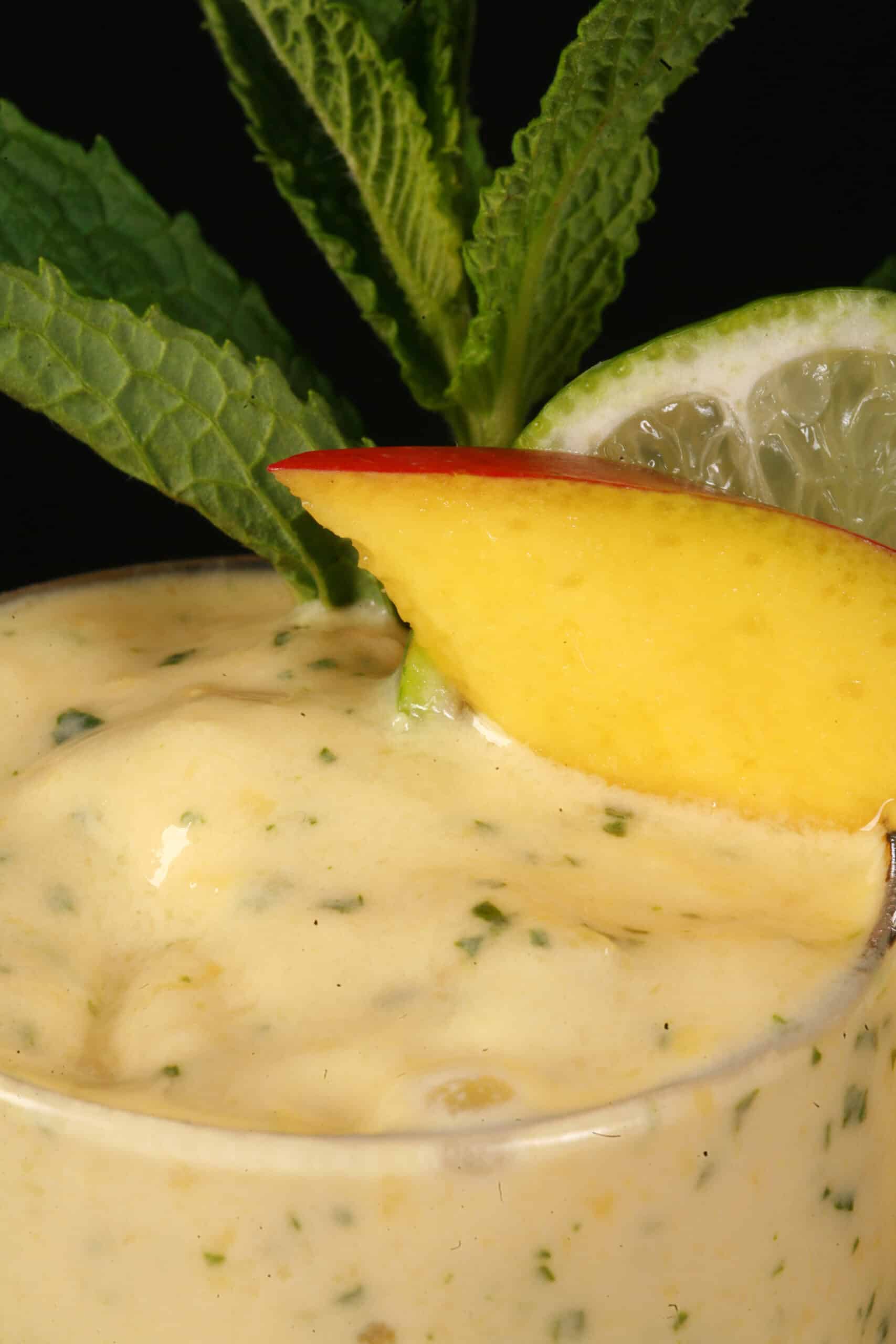 A high protein mango mojito smoothie, garnished with fresh mango and lime slices and a mint sprig.