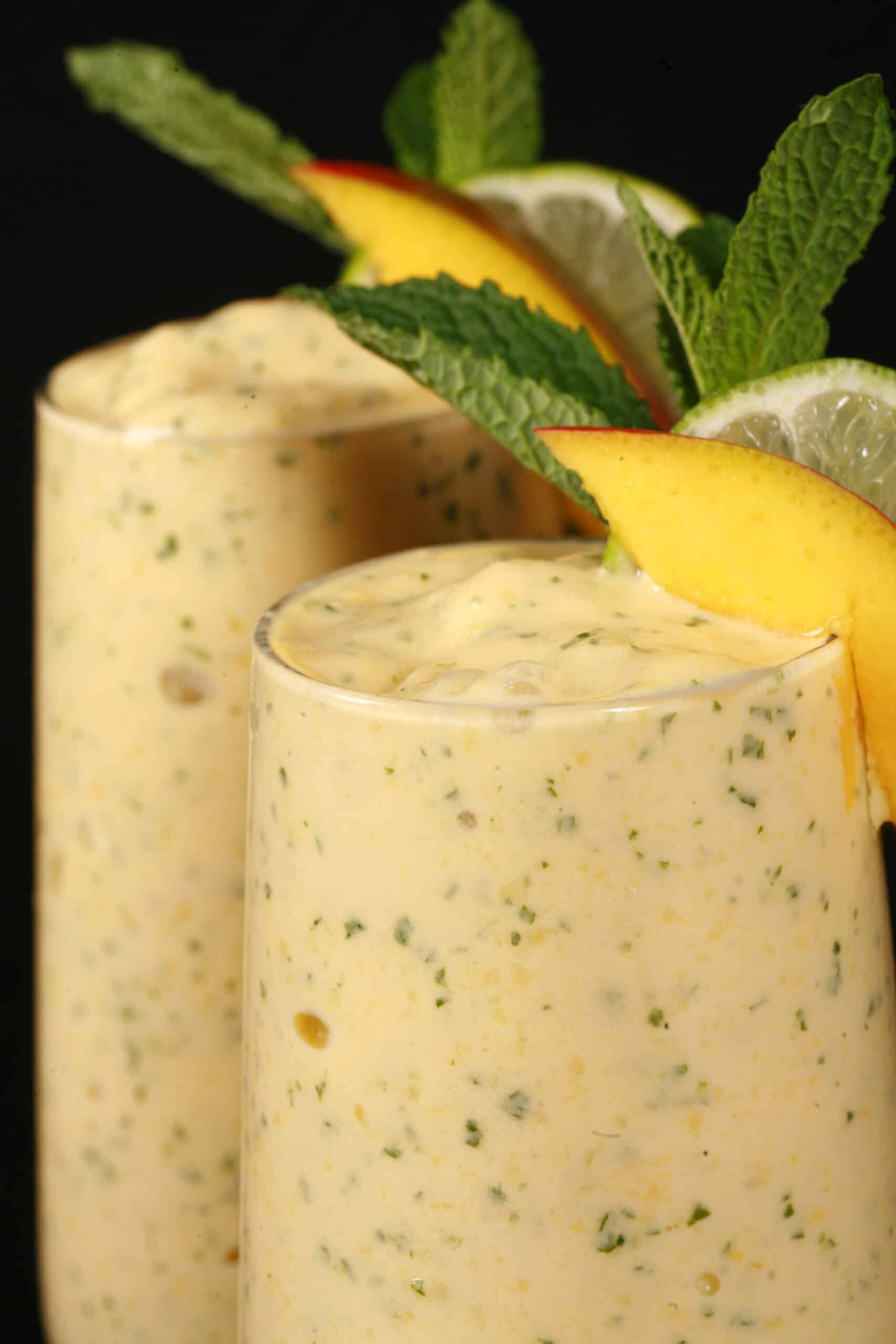 2 high protein mango mojito smoothies, garnished with fresh mango and lime slices and a mint sprig.