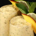 2 high protein mango mojito smoothies, garnished with fresh mango and lime slices and a mint sprig.