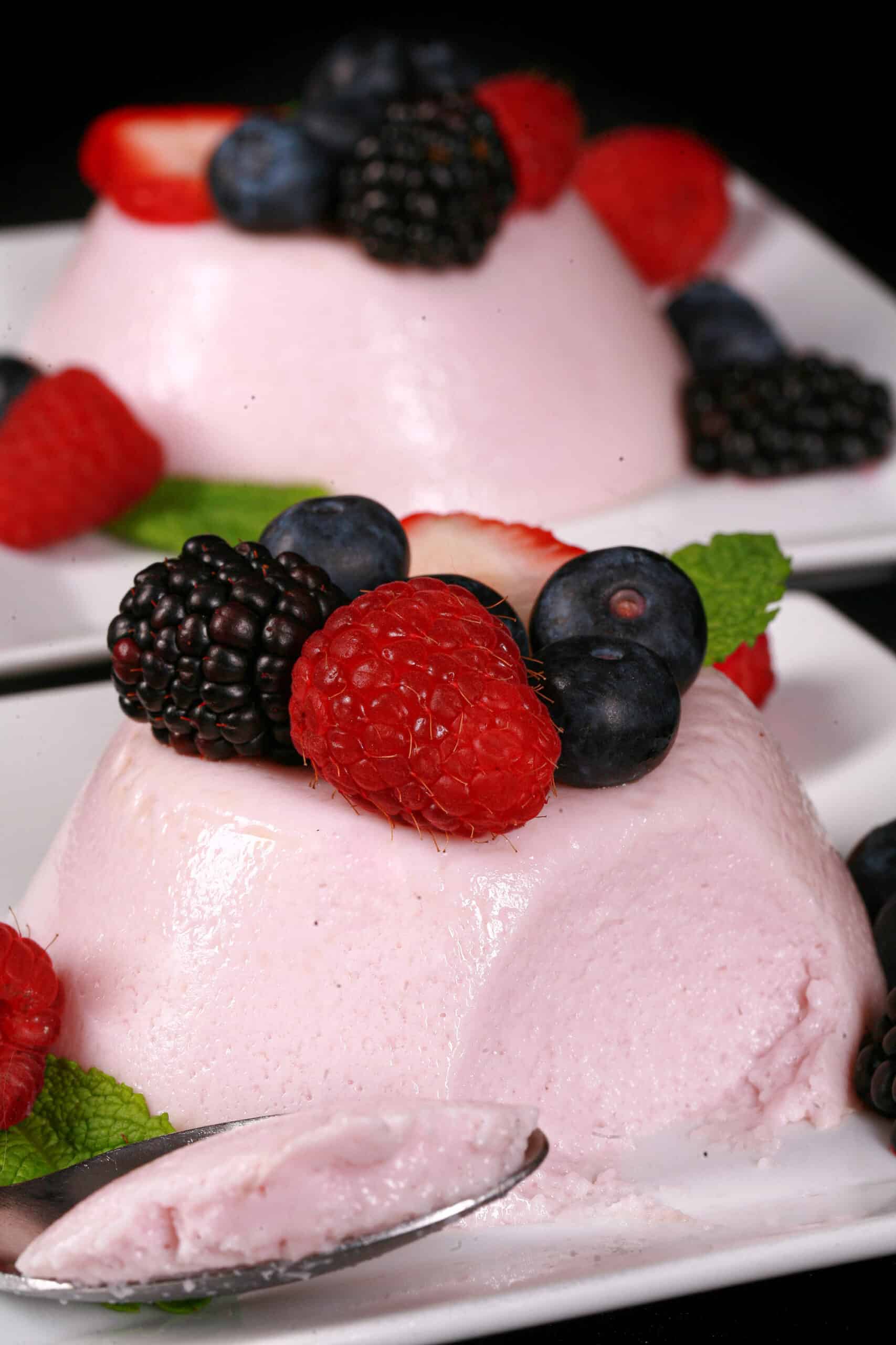 2 light pink high protein panna cotta servings on plates, with mixed berries and mint leaves.