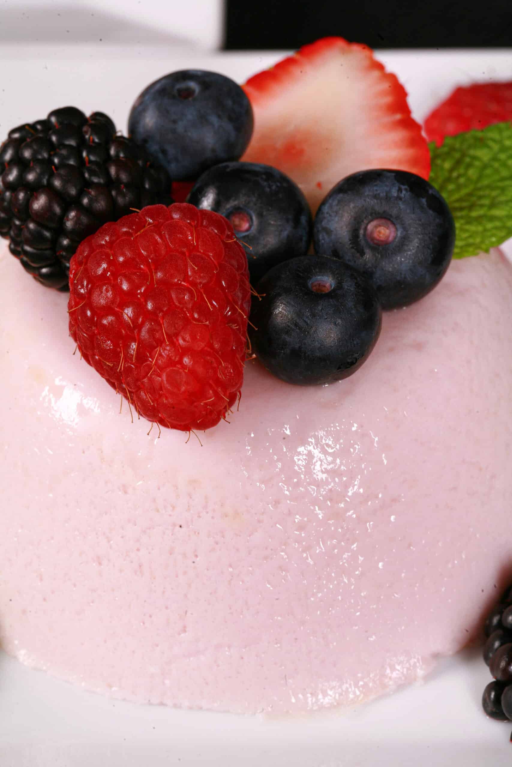 A light pink protein powder panna cotta on a plate, with mixed berries and mint leaves.