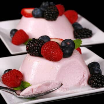 2 light pink high protein panna cotta servings on plates, with mixed berries and mint leaves.