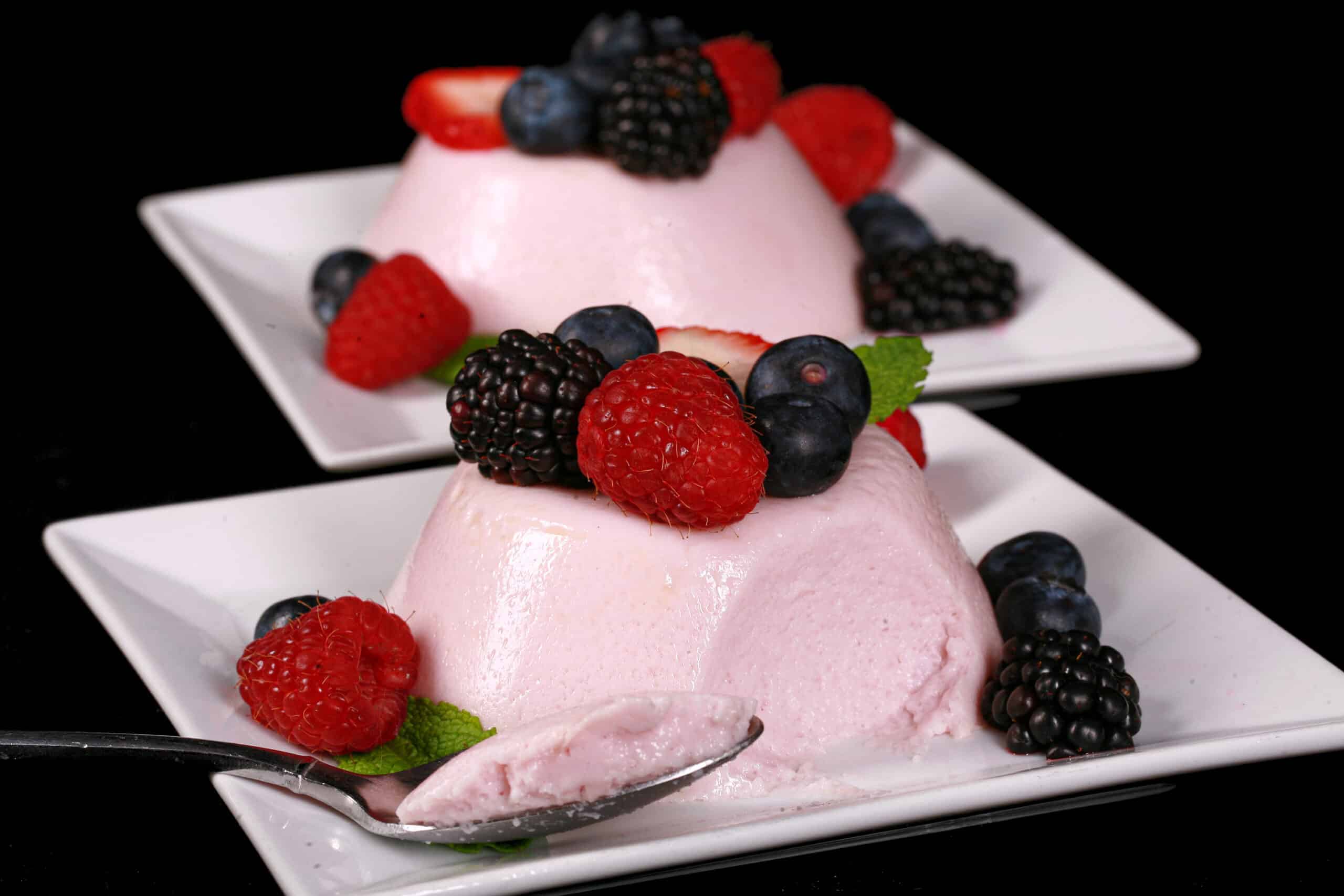 2 light pink high protein panna cotta servings on plates, with mixed berries and mint leaves.