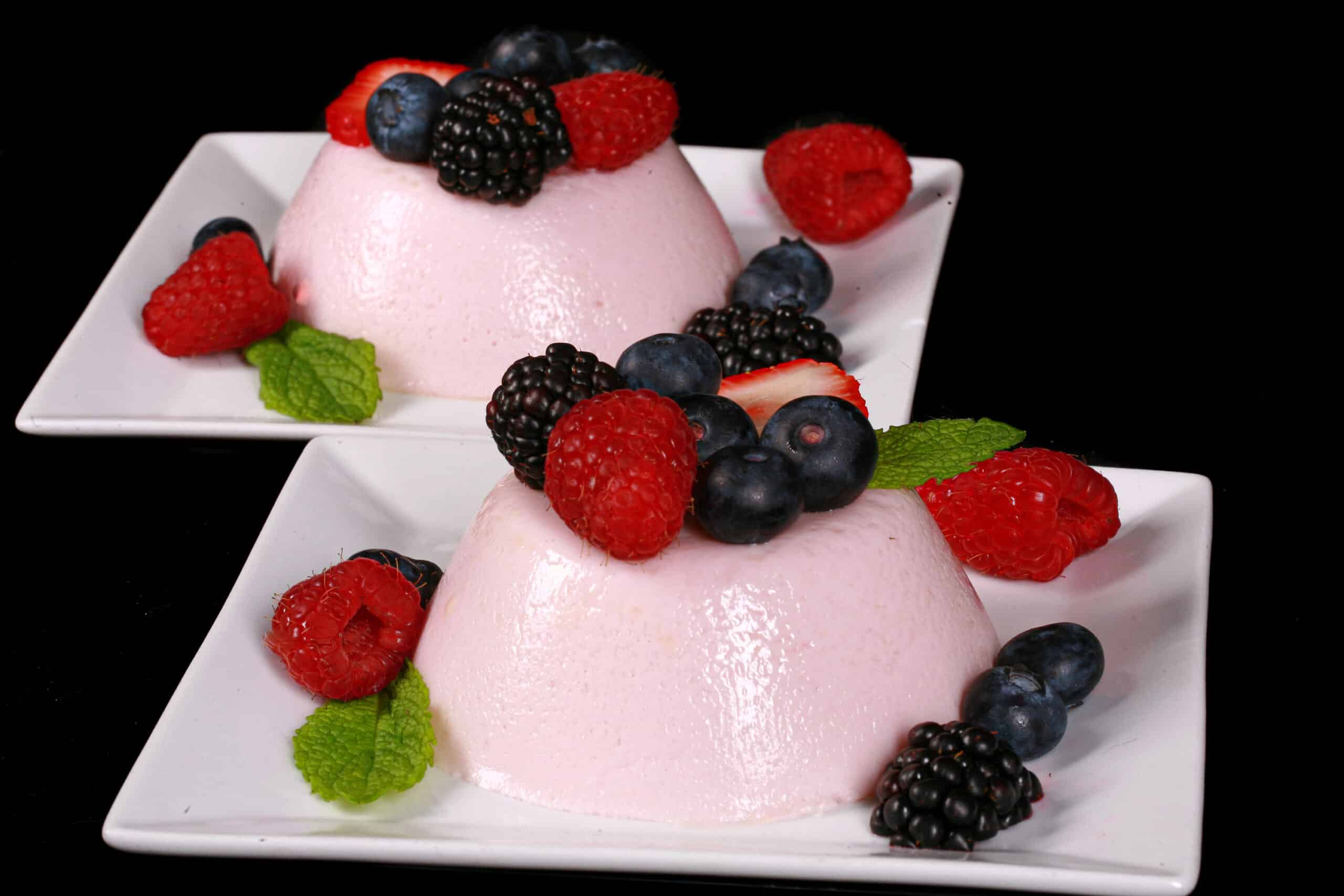 2 light pink protein powder panna cotta servings on plates, with mixed berries and mint leaves.