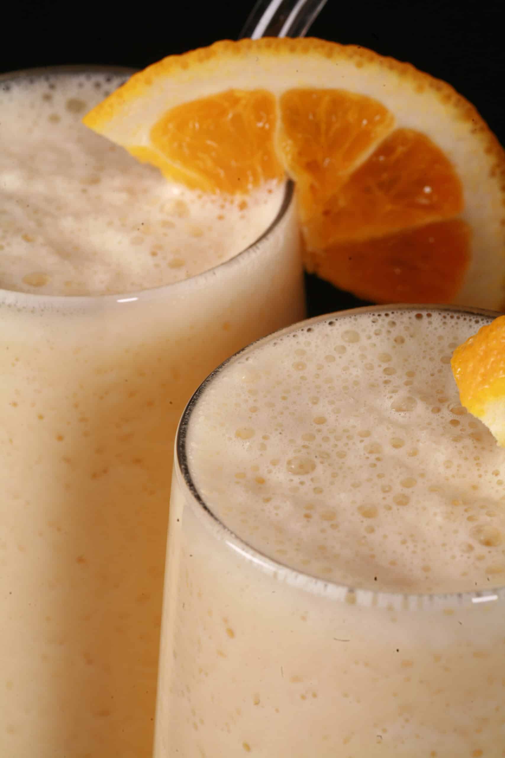 Two tall glasses of protein powder Orange Julius smoothies, garnished with fresh orange slices.