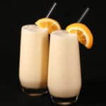 Two tall glasses of protein powder Orange Julius smoothies, garnished with fresh orange slices.
