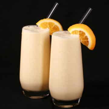 Two tall glasses of protein powder Orange Julius smoothies, garnished with fresh orange slices.