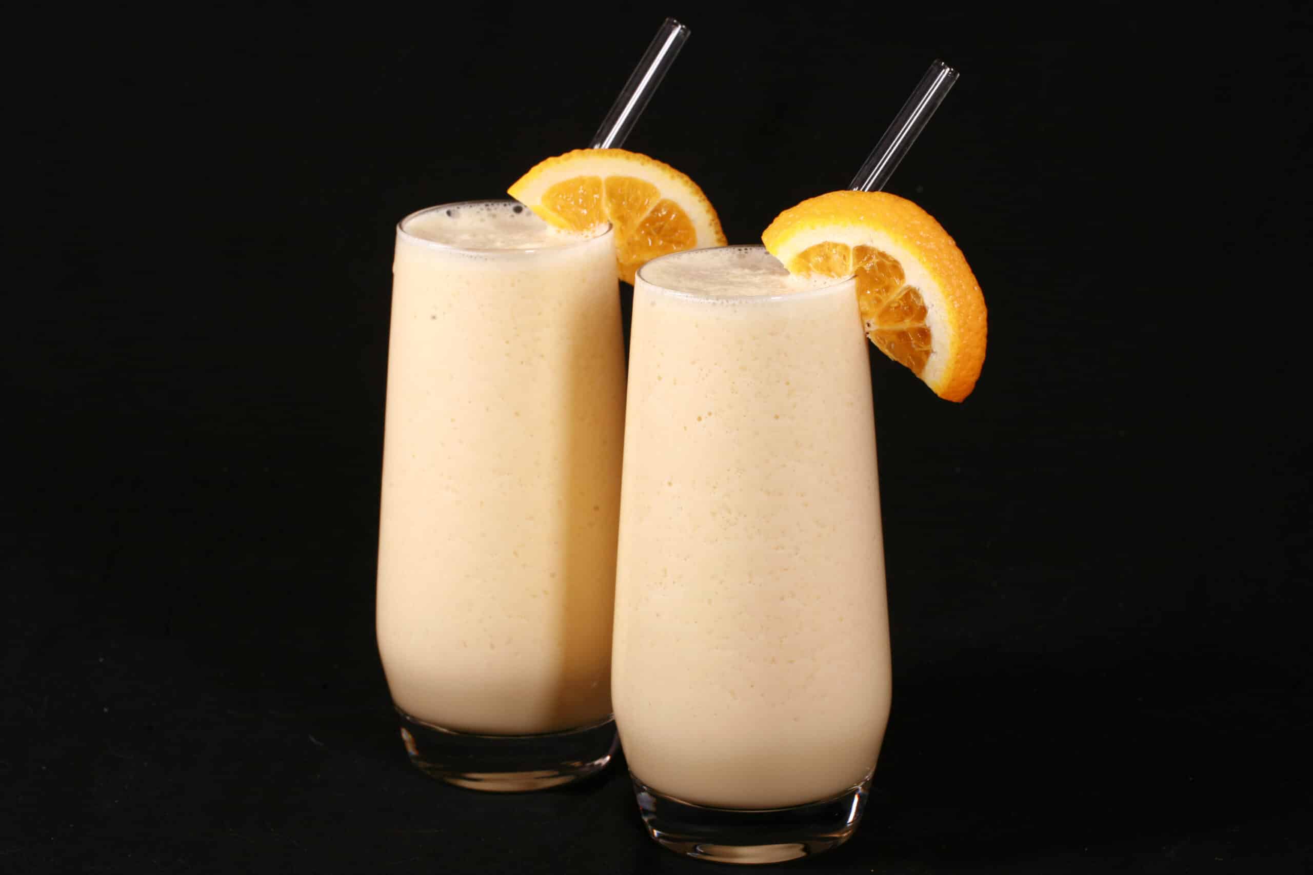 Two tall glasses of protein powder Orange Julius smoothies, garnished with fresh orange slices.